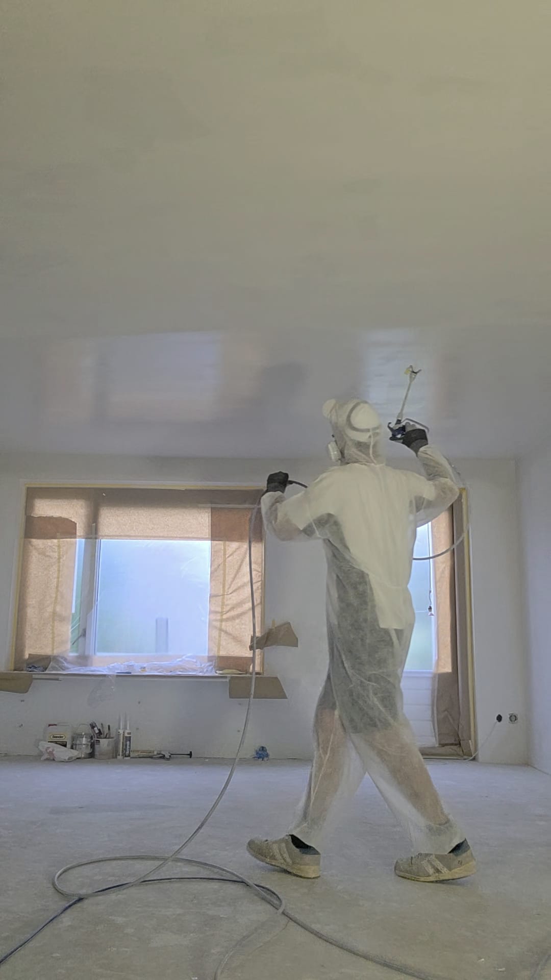 Stukadoor in witte overall spuit een plafond in een afgeplakte kamer