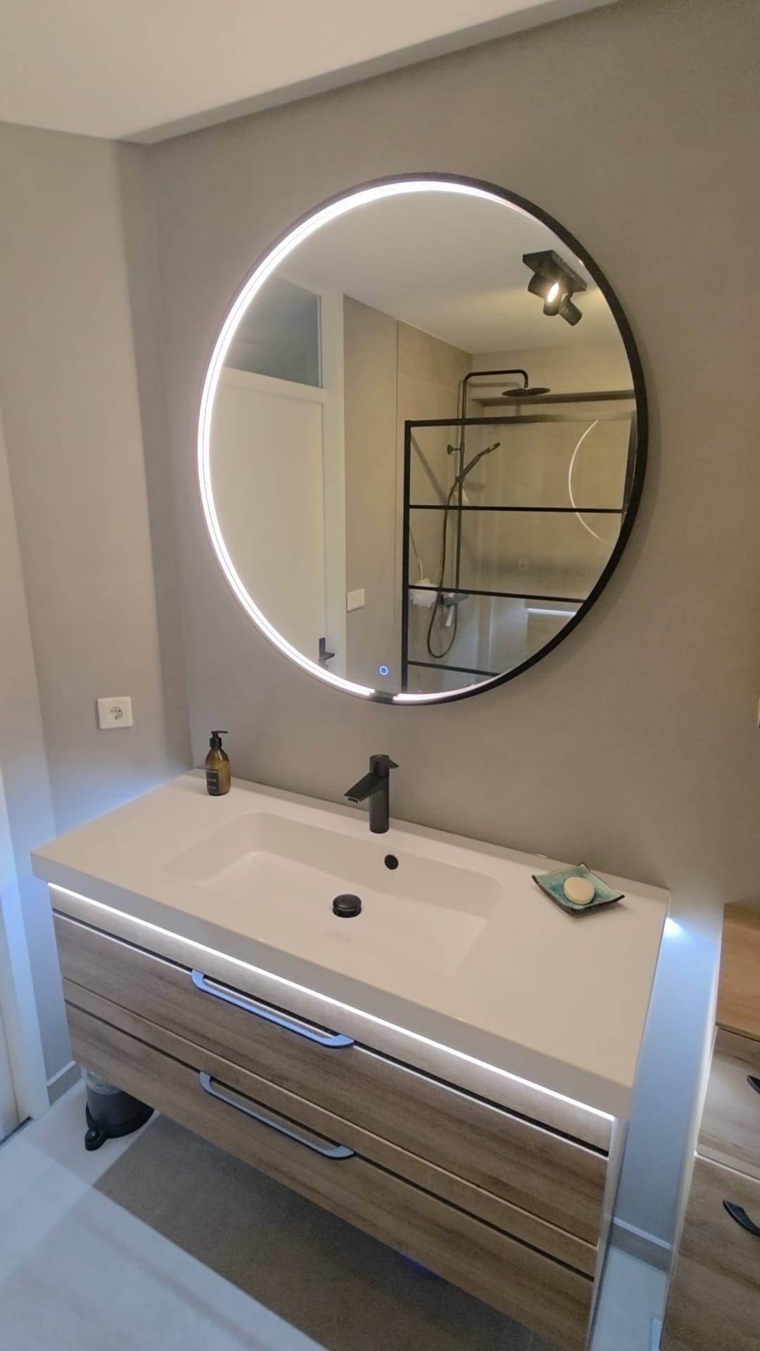 Badkamer met grijze betonlook wanden, ronde verlichte spiegel en houten meubel